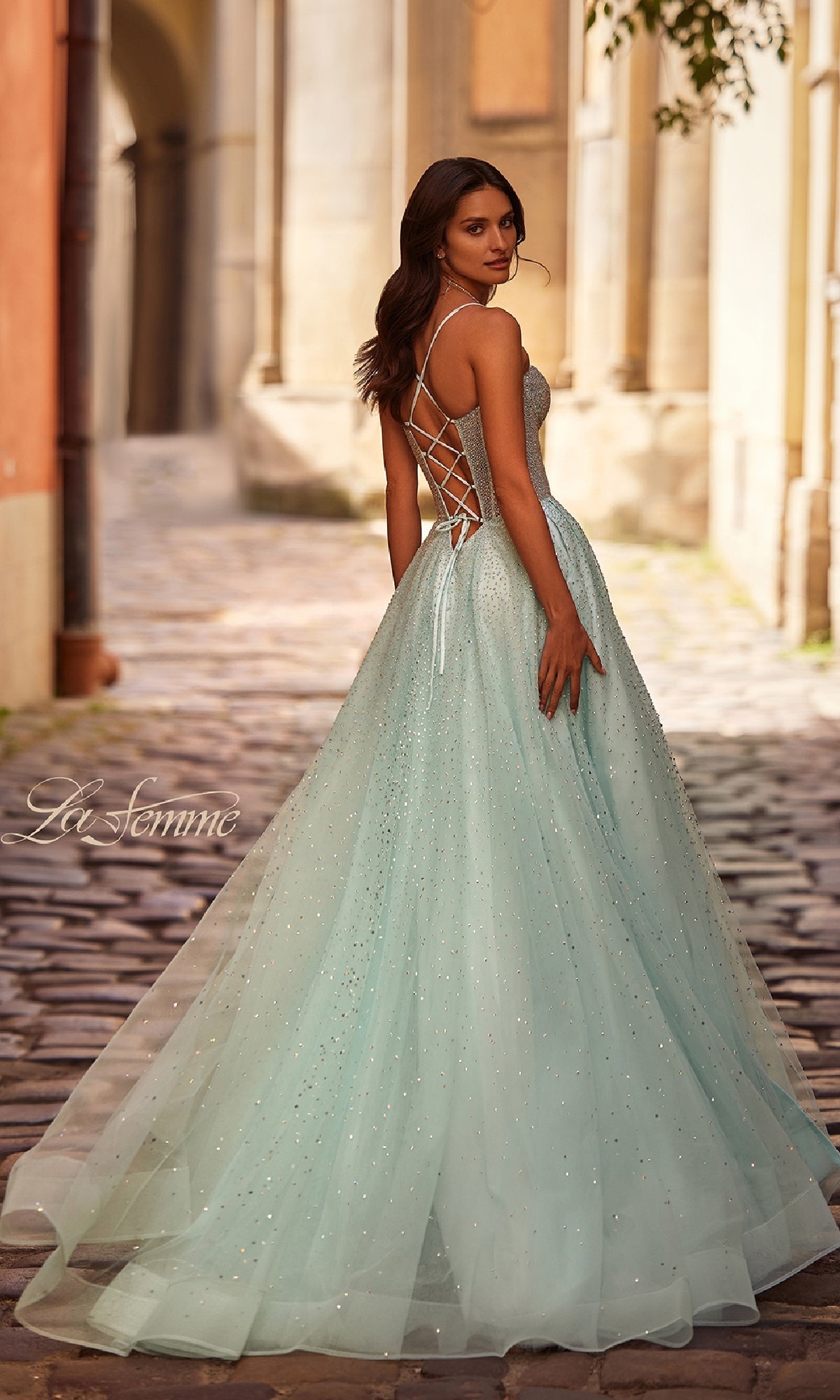 A woman in the Lace-Up Beaded Long Prom Ball Gown: La Femme 32725 stands on a cobblestone street, looking over her shoulder in the sparkling blue dress with a lace-up back.